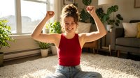 Young Caucasian woman celebrating success with raised fists in bright living room. Happy female showing victory gesture at home with plants and natural light.