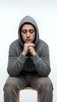 Depressed middle-aged Caucasian man in gray hoodie sitting on chair with closed eyes and clasped hands showing mental health struggles and sadness.