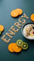 Word ENERGY made from oats and seeds beside a yogurt bowl with granola, kiwi and persimmon slices on teal background, healthy breakfast concept.