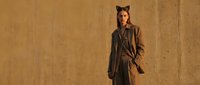 Young Caucasian woman in oversized brown coat with cat ears headband poses confidently against warm textured concrete wall in autumn fashion editorial.