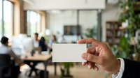 Close-up of hand holding blank business card in modern office environment with colleagues working at desk in background, suitable for corporate branding and networking concepts.