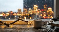 Cozy winter evening scene with warm coffee mug, fairy lights and city bokeh through rainy window creating peaceful hygge atmosphere for relaxation.