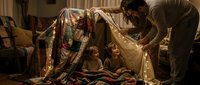 Father building cozy blanket fort with two young children in warm living room, creating magical family bonding moment during evening playtime at home.
