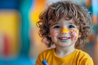 Cheerful child with curly hair wearing yellow t-shirt and colorful face paint butterfly design smiling at camera against blurred festive background.