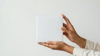 Woman with medium skin tone wearing a white long-sleeve shirt holding and presenting a blank white square card or product box against neutral background for mockup and branding purposes.