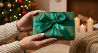 Woman hands holding elegant green gift box with satin ribbon bow in cozy Christmas living room with decorated tree and warm candles creating festive atmosphere.