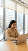 Young woman of South Asian descent working at laptop in modern high-rise office with city skyline view, wearing beige sweater, suitable for business, technology, and corporate concepts.