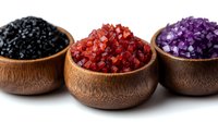 Three wooden bowls filled with colorful sea salt crystals in black, red, and purple arranged on white background for culinary and spa applications.