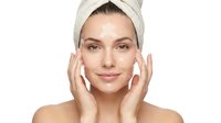 Young woman applying facial cream with towel on head after shower on white background for skincare routine and beauty treatment advertising.