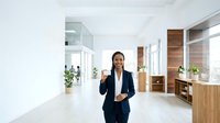 Professional woman with dark hair and medium complexion wearing navy blazer standing in modern office hallway with wooden accents, potted plants, and natural light for corporate business concepts.