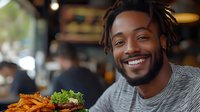 Young African American man with dreadlocks and beard smiling warmly while enjoying burger and french fries at casual restaurant, genuine happiness during dining experience.