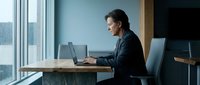 Young professional man of South Asian descent working on laptop at wooden desk in modern office with city skyline view, wearing dark blazer, suitable for business, technology, and corporate concepts.