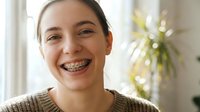 Young Caucasian teenage girl with metal braces smiling happily at camera in bright home interior, orthodontic dental treatment and oral health care concept.