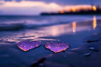 Two purple heart-shaped objects on wet beach sand at sunset with blurred city lights reflecting on water creating romantic coastal atmosphere.