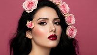 Young Asian woman with dark hair adorned with pink roses, wearing bold red lipstick and glamorous makeup against pink background for beauty and fashion concepts.