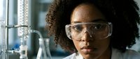 Young female scientist with curly dark hair and medium brown skin tone wearing clear safety glasses, conducting laboratory research in a modern scientific facility with glass equipment and blurred background.