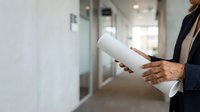 Professional woman holding rolled blueprints in modern office hallway, wearing dark blazer. Architectural plans for construction, engineering, and business project planning.