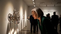 Mature woman with gray hair examining contemporary art sculpture in modern gallery space with visitors in background.