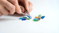 Hands holding tweezers picking up colorful plastic pieces for crafting, sorting, or educational activities on white background.