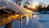 Icicles hanging from wooden railing at sunset, golden light illuminating frozen water droplets against snowy winter landscape.
