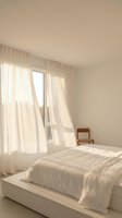 Minimalist bedroom interior with flowing white curtains and natural sunlight creating warm shadows on neutral walls and bedding.