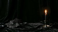 Ornate silver crown rests on dark fabric beside burning candle in candlestick, creating moody, dramatic lighting for fantasy, medieval, or gothic themed designs and storytelling projects.