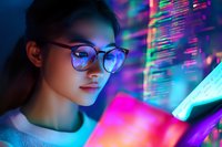Young Asian female programmer in glasses illuminated by colorful neon light from multiple screens, looking at glowing code display in dark environment.