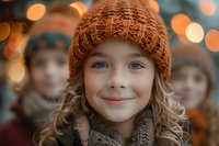 Young girl wearing knitted orange beanie and scarf smiles warmly at camera with bright blue eyes and rosy cheeks against soft bokeh holiday lights background.