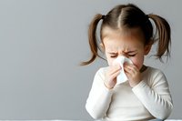 Young Caucasian girl with pigtails wearing white sweater sneezing into tissue, showing cold and flu symptoms against light gray background. For health and medical concepts.