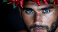 Close up portrait of young bearded man with intense blue eyes wearing red chili pepper crown against dark background, dramatic lighting highlights facial features.