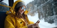 Woman in bright yellow winter jacket and knit hat using smartphone on train journey through snowy mountain landscape.