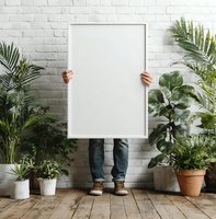 Person holding blank white poster frame surrounded by potted plants against white brick wall, perfect for mockup designs.