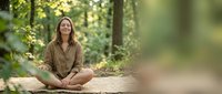 Young Caucasian woman meditating in peaceful forest setting, sitting cross-legged on wooden platform surrounded by lush green trees and natural sunlight.