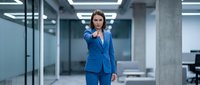 Professional businesswoman in blue suit pointing forward in modern office environment with glass walls and contemporary lighting fixtures.