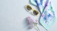 Stylish summer flat lay with yellow sunglasses, floral silk scarf and lilac branch on light background for fashion and lifestyle content.