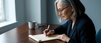 Senior businesswoman with gray hair and glasses writing in notebook at wooden desk with coffee cup by window in modern office setting.
