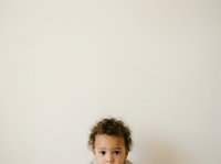 Young child with curly brown hair and light brown skin looking at camera against neutral beige wall background, suitable for parenting, childcare, education, and family-related content.