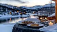 Romantic winter evening with wine glasses and dessert plates on balcony overlooking snowy mountain lake at dusk, candlelit dining atmosphere.
