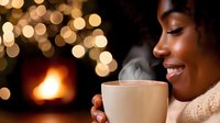 Young African American woman enjoying hot beverage near fireplace with bokeh lights in background, expressing joy and contentment in cozy winter atmosphere.