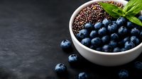 Fresh ripe blueberries and black quinoa in white ceramic bowl on dark background with mint leaves, healthy superfood breakfast ingredients.