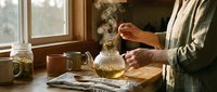 Woman preparing herbal tea in cozy kitchen with glass teapot and dried herbs on wooden table near window creating warm morning atmosphere.