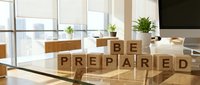 Young professional woman with light skin tone in modern office environment arranging wooden blocks spelling "Be Prepared" on desk, symbolizing business readiness and planning strategy.