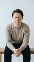 Young woman experiencing stress and anxiety sitting on bench with eyes closed and worried expression in beige sweater against white background.