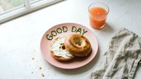 Bagel with cream cheese on a pink plate with Good Day lettering, served with a glass of grapefruit juice for a cheerful morning breakfast.