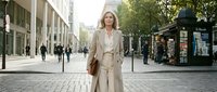 Professional blonde businesswoman in beige suit walking confidently on urban street with modern buildings and cobblestone pavement for corporate lifestyle content.