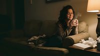 Young woman with long dark wavy hair and olive complexion sitting on beige couch holding tissue box, appearing emotional or unwell during indoor moment.