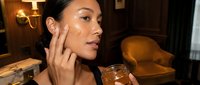 Young Asian woman applying natural skincare cream from glass jar in cozy bedroom setting with warm lighting for beauty routine and self-care wellness.