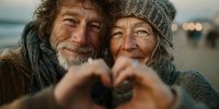 Happy senior couple making heart shape with hands outdoors, showing love and affection in golden hour light with warm smiles and winter clothing.