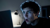 Shocked man with dark hair staring at laptop screen in dimly lit room, expressing surprise or concern while working late at night on computer.
