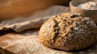 Freshly baked whole grain bread loaf with seeds on wooden surface, flour dusted cloth nearby. Artisan bakery, food photography, culinary content.